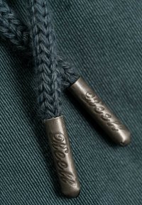 Cordones de zapatos trenzados de color azul oscuro con terminales de metal grabados con "reell," sobre un fondo de tela texturizada.