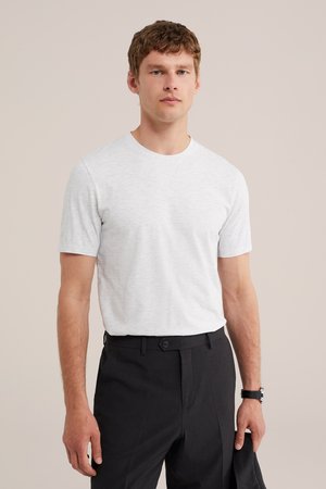 Man draagt lichtgrijs T-shirt met korte mouwen en zwarte broek, houdt zwarte jas vast, staat voor een beige achtergrond.