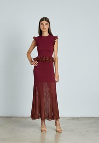 Donna in piedi che indossa un abito senza maniche color bordeaux con dettagli a volant, una gonna plissettata trasparente e sandali open-toe beige su uno sfondo neutro.