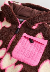 Veste en polaire avec motifs de cœurs marron et crème, fermeture éclair rose et poche poitrine matelassée rose avec tirette noire.