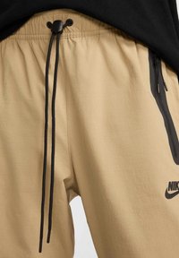 Beżowe spodnie sportowe z elastycznym pasem, czarnym sznurkiem i bocznymi kieszeniami na zamek. Z przodu znajduje się czarne logo Nike.