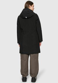Frau in einer schwarzen Kapuzenparka-Jacke und lockeren braunen Cargo-Hosen, die mit dem Rücken zu einer hellen, einfarbigen Hintergrund steht.
