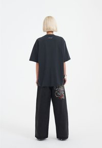 Černé oversized tričko s krátkými rukávy, na zadní straně má decentní logo; v kombinaci s černými širokými džíny zdobenými barevnou výšivkou draka.