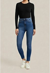 Modré džínové kalhoty s vysokým pasem a úzkým střihem, s kapsami na zadní straně, doplněné těsným černým dlouhým rukávem a bílými teniskami.