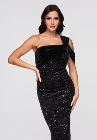 Czarna wieczorowa suknia z aksamitnym gorsetem i spódnicą ozdobioną cekinami. Asymetryczny krój z dekoracyjnymi marszczeniami na materiale na jednym ramieniu.