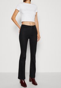 Jeans negros de campana combinados con una camiseta ajustada blanca con bordado sutil. El modelo lleva botas de tacón cuadrado burdeos.