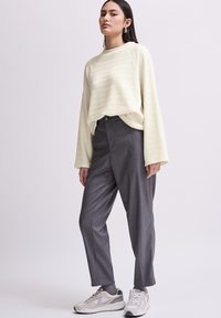 Beige strukturierter Sweatshirt mit langen Ärmeln, kombiniert mit grauen maßgeschneiderten Hosen und hellgrauen Sportschuhen. Das Modell steht in einem neutralen Umfeld.