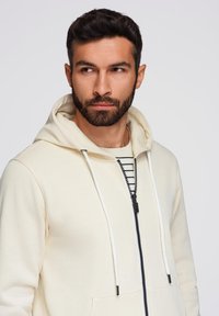 Felpa zip con cappuccio di colore crema chiaro, dotata di tasca a marsupio e cordini bianchi. Indossata sopra una maglietta a righe bianco e nero, con una texture liscia.