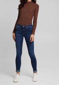 Top de manga larga marrón con canalé, combinado con vaqueros ajustados de color azul oscuro. Los vaqueros tienen un diseño clásico de cinco bolsillos y los puños doblados. Zapatillas blancas.