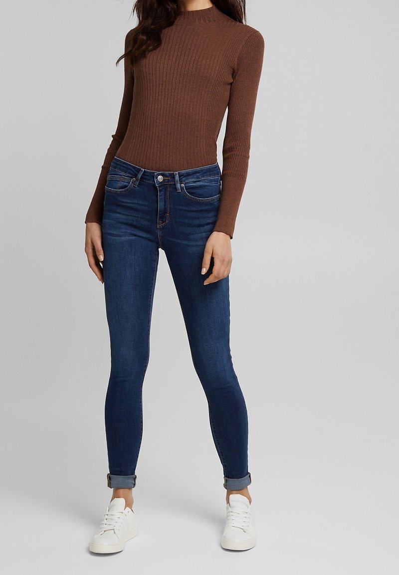 Top de manga larga marrón con canalé, combinado con vaqueros ajustados de color azul oscuro. Los vaqueros tienen un diseño clásico de cinco bolsillos y los puños doblados. Zapatillas blancas.