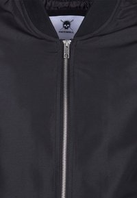 Veste noire avec une fermeture éclair argentée et un col côtelé, portant une étiquette "Tuffskull" avec un logo de crâne et d'aiguilles à tricoter croisées.