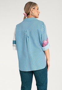 Koszula w niebiesko-białe paski o luźnym kroju, z kwiatowym haftem na rękawach oraz plisą z tyłu dla ułatwienia ruchu.