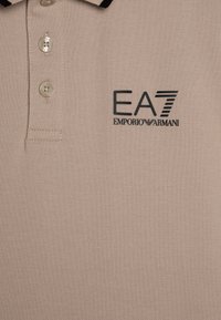 Beige polo shirt laget av strukturert stoff, med tre knapper, svart kragekant og EA7 Emporio Armani-logo i mørk trykk.