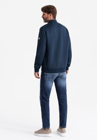 Veste bleu marine avec col montant, en tissu texturé, et écusson sur la manche, associée à un jean bleu clair et des baskets beiges.