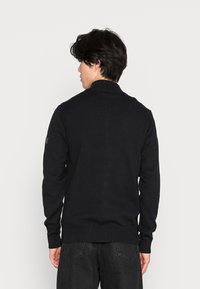 Zwarte cardigan met een hoge kraag, geribbelde manchetten en zoom. Beschikt over een subtiele textuur en een klein logopatch op de linkermouw.