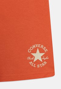 Oranžové bavlněné tričko s krémovým kulatým logem, na kterém je hvězda a nápisy "Converse All Star" a "Chuck Taylor."