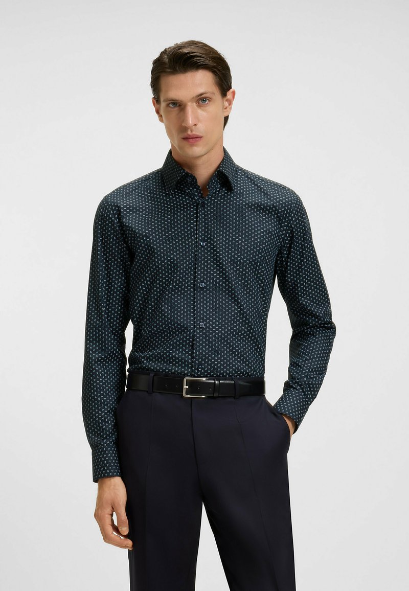 Tummansininen nappikaulustakki, jossa on pieni valkoinen geometrinen kuvio, pitkät hihat ja terävä kaulus. Yhdistettynä mustiin housuihin ja vyöhön.