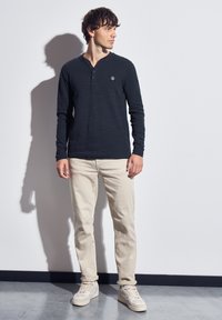 Long-sleeve navy henley shirt with a small logo patch, paired with beige slim-fit pants and light-colored sneakers, against a plain background.