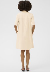 Svetlo béžové šaty košeľového strihu s krátkymi rukávmi, golierom, detailom na chrbte s plisovaním a bočnými vreckami. Nosiť s čiernymi plochými sandálmi.