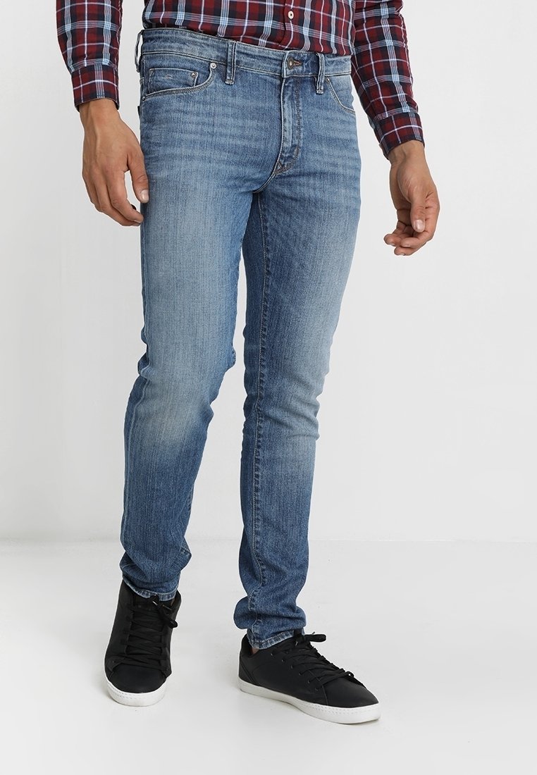 Dark blue denim jeans with a slim fit, lightly distressed texture, and five-pocket design, paired with black sneakers.