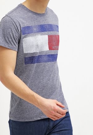 Mann trägt ein hellgraues T-Shirt mit großem rechteckigem rot-weiß-blauem Motiv und dunkelblauen Jeans, dargestellt von Hals bis Hüfte.
