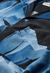 Tissu à motif abstrait bleu et noir avec une poche avant et des boutons noirs sur un vêtement étiqueté "MINIM".