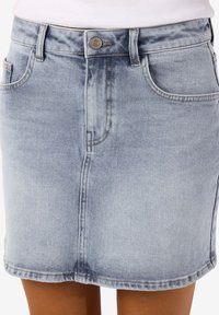 Φούστα από ανοιχτό γαλάζιο τζιν με κουμπιά στο μπροστινό μέρος και τσέπες, φορεμένη με λευκό τοπ, που φτάνει μέχρι το μέσο του μηρού και έχει εφαρμοστή γραμμή.