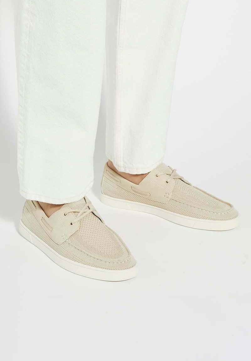 Baskets en maille beige avec semelles en caoutchouc blanc, présentant des lacets et des détails de couture. Portées avec un pantalon blanc ample.