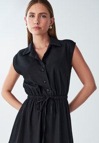 Czarna sukienka bez rękawów z zapinaniem na guziki z przodu, kołnierzykiem oraz ściąganym w pasie sznurkiem. Miękka tkanina o gładkiej fakturze.