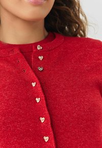 Red cardigan made of textured fabric, featuring gold heart-shaped buttons down the front. Round neckline with subtle ribbing.