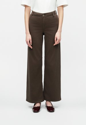 Femme portant un pantalon large taille haute marron foncé avec un bouton avant et une fermeture éclair, associé à des ballerines en cuir verni bordeaux.