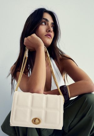Young woman with long dark hair holds cream-colored quilted purse with gold chain strap, wearing white top and dark green pants.