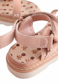 Sandale en daim rose avec semelle intérieure à motifs floraux, brides réglables et épaisse semelle crème. Présente des détails de couture autour du bord de la semelle.