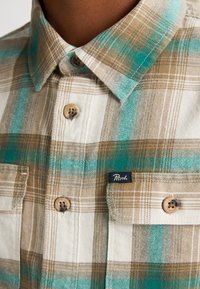 Košile s límečkem v béžovém, bílém a tyrkysovém kostkovaném vzoru, s béžovými knoflíky a malou nášivkou "Petrol" na náprsní kapse.