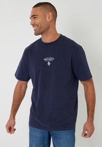 Hombre sonriendo de lado, vistiendo una camiseta gráfica azul marino con la palabra "Provence" y jeans azules, sobre un fondo blanco liso.