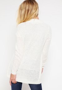 Frau mit langen blonden Haaren trägt einen lockeren, weißen Strickpullover und dunkelblaue Jeans, von hinten vor einem weißen Hintergrund gezeigt.