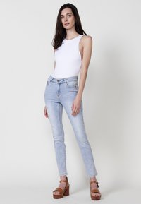 Bodysuit blanco con cortes en los laterales, jeans de mezclilla azul claro con sutiles adornos y sandalias de tacón de madera marrón.