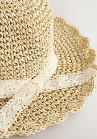 Chapeau en paille tressée avec un large bord festonné et un léger ruban en dentelle crème noué autour de la couronne. Couleur beige naturel et finition texturée.