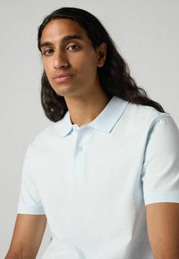 Ljusblå polo skjorta gjord av mjukt tyg, med klassisk krage, tre knappar och korta ärmar. Slät textur med subtil märkeslogotyp.