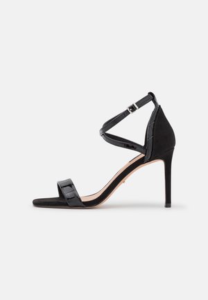 Sort højhælet sandal med åben tå, ankelrem med spænde og en stiletthæl på en ensartet lys baggrund.