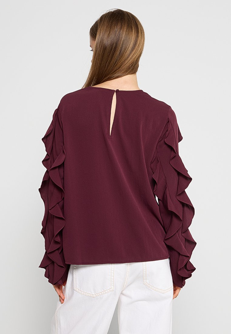 Bordeaux blouse met ruffled lange mouwen, gladde textuur, ronde halslijn en een klein keyhole detail aan de achterkant. Gepaard met witte jeans.