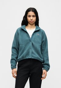 Γυναίκα με μακριά σκουρόχρωμα μαλλιά, που φοράει πετρόλ φλις ζακέτα πάνω από ένα λευκό πουκάμισο και μαύρο παντελόνι, στέκεται μπροστά σε ένα απλό φωτεινό φόντο.