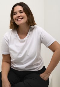 Bílý krátký rukáv tričko s kulatým výstřihem, vyrobené z hladké tkaniny, nošené s černými kalhotami. Jednoduchý design, žádné viditelné vzory ani loga.