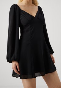 Kvinna klädd i en svart klänning med långa ärmar, djup V-ringning och puffärmar, stående mot en enkel ljus bakgrund.