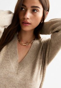 Beige gebreide trui met V-hals en geribde manchetten, gecombineerd met een gouden en bruine schakelketting. Smooth textuur, minimaal patroon.