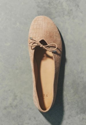 Béžové semišové baleríny s reliéfnou mriežkovanou textúrou a detailom prednej mašle, zobrazené zhora na sivom povrchu.