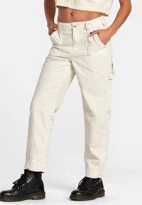 Crème-kleurige cargobroek met een ontspannen pasvorm, meerdere zakken en een knoopsluiting. Gestyled met zwarte veterschoenen.