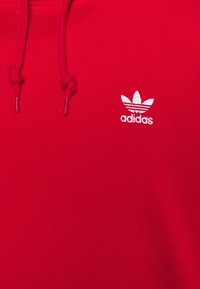 Röd hoodie i texturerat tyg med justerbara snören. Har en liten vit Adidas-logotyp på vänstra bröstet.