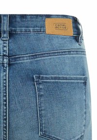 Jasnoniebieska tylna kieszeń dżinsów z denimu z jasnobrązową skórzaną metką z nazwą marki „camel active” przeszytą na pasku od spodni.