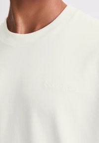 Helles graues Sweatshirt mit geripptem Rundhalsausschnitt und geprägtem Logodetail. Weiche Textur und minimalistisches Design.